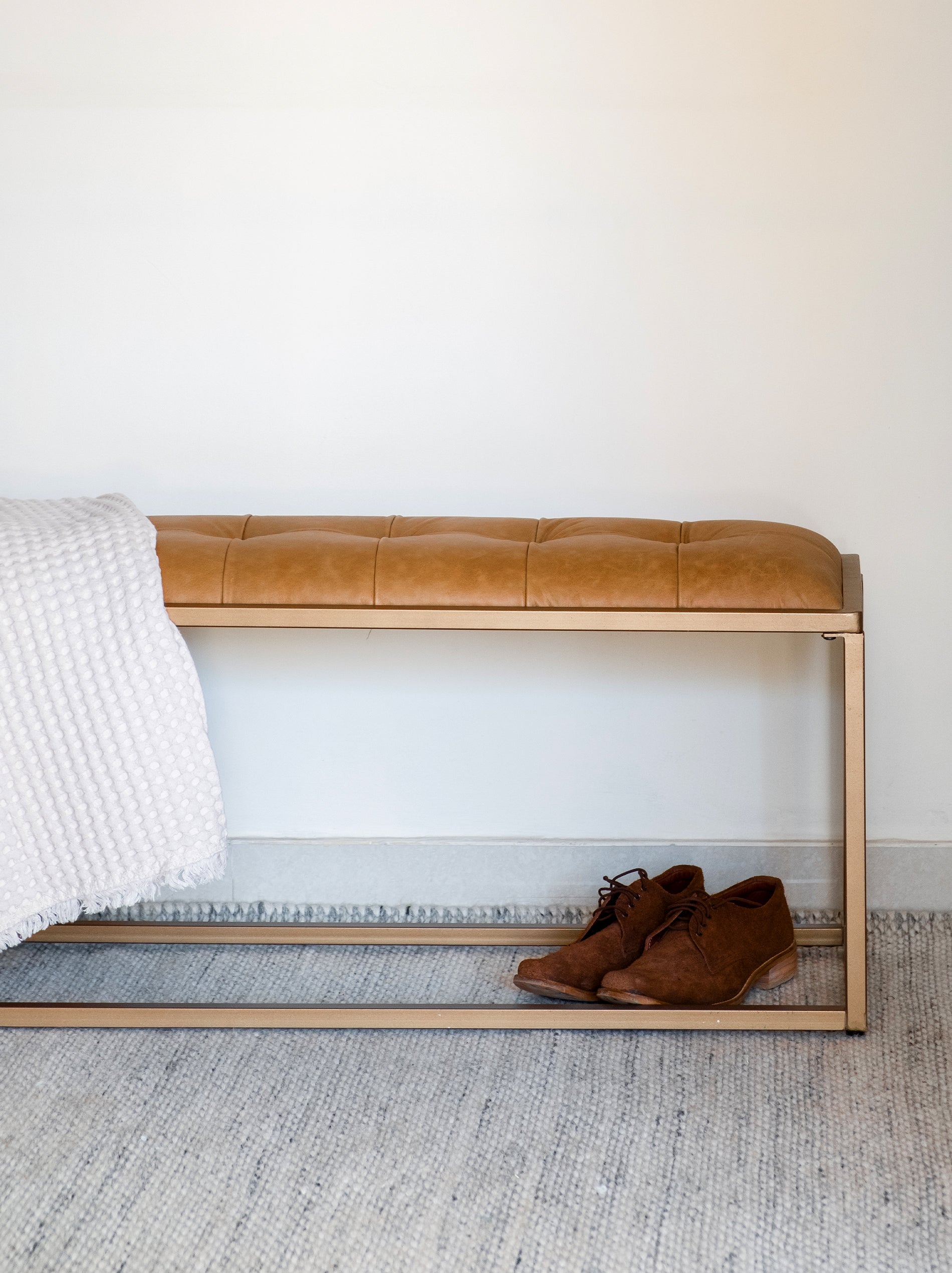 An upholstered bench with brown leather seating and a golden iron frame, placed at the foot of a white bed with a grey blanket, next to a pair of brown shoes.