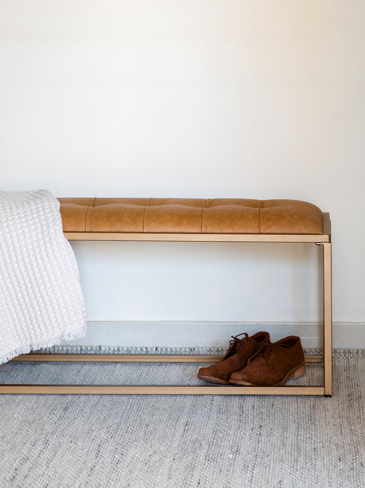 An upholstered bench with brown leather seating and a golden iron frame, placed at the foot of a white bed with a grey blanket, next to a pair of brown shoes.