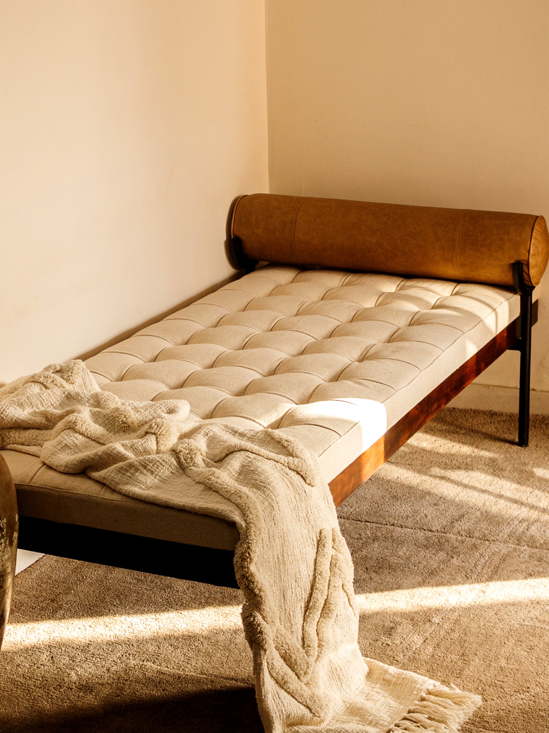 A day bed with a beige cotton cushion on a mango wood frame with iron legs, topped with a brown faux leather pillow.