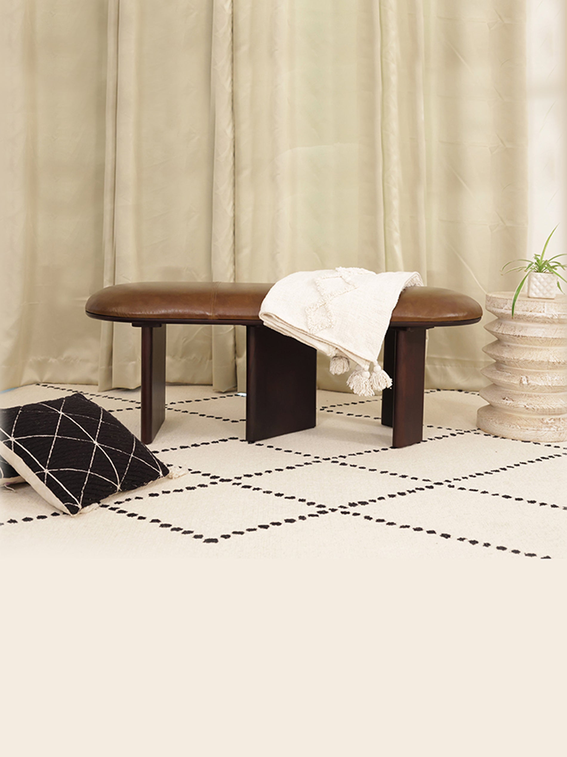 Wooden bench with a white blanket draped over it in a room with beige curtains and a black and white patterned rug.